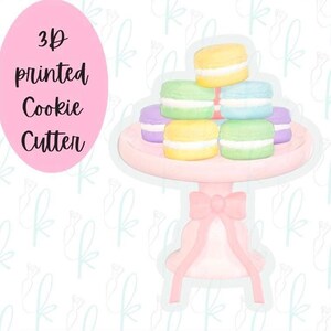 Puede incluir: Un cortador de galletas impreso en 3D con forma de soporte de postre escalonado con macarons coloridos. Los macarons son amarillos, verdes, azules y morados con relleno blanco. El soporte es rosa con un lazo.
