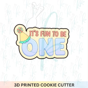 May include: A 3D printed cookie cutter in the shape of the phrase "It's Fun to Be One." The design features a party hat and the word "ONE" in blue, with the text "It's Fun to Be" in pink. The cutter has a yellow border.