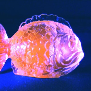 May include: A decorative glass fish figurine in shades of orange and pink, with intricate details. The fish has a textured surface and is set against a dark blue background. The figurine is a unique home decor item.
