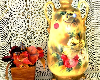 May include: An antique vase with a floral design and gold detailing, next to a small wooden planter with artificial flowers. The vase has a cream base with hand-painted red, yellow, and white flowers. The background is a white lace doily.