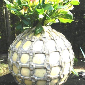 May include: A decorative, spherical vase with a woven metal design, filled with green leaves and yellow and pink flowers. The vase has a light yellow base color and sits on a dark surface.