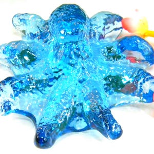 Könnte beinhalten: Eine durchscheinende, blaue Glas-Oktopus-Figur mit acht strukturierten Armen. Der Oktopus hat eine unebene, blasenartige Oberfläche, und die Farbe variiert von hell- bis tiefblau. Die Figur ist ein dekorativer Artikel.