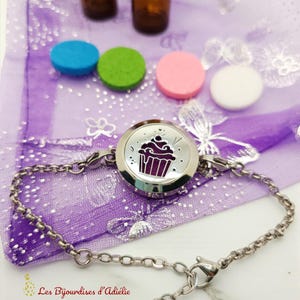 May include: A silver bracelet with a circular pendant displaying a purple cupcake design. The image includes small, colourful, round diffusers in blue, green, pink, and white, and two small brown bottles. The background is a sheer purple fabric.