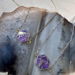 May include: Two necklaces with hexagonal pendants. One necklace has a gold chain and pendant, the other has a silver chain and pendant. The pendants are purple with gold and silver accents. The necklaces are displayed on a light-colored, patterned surface.