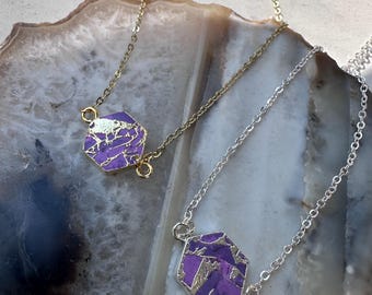 May include: Two necklaces with hexagonal pendants. One necklace has a gold chain and pendant, the other has a silver chain and pendant. The pendants are purple with gold and silver accents. The necklaces are displayed on a light-colored, patterned surface.