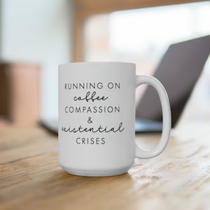 May include: White ceramic coffee mug with black text that reads "Running on coffee, compassion & existential crises".