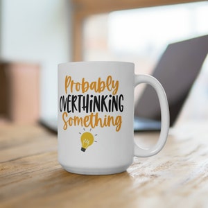 May include: White ceramic mug with the text "Probably Overthinking Something" in black and orange lettering. A yellow lightbulb graphic is below the text. The mug has a large handle and is sitting on a wooden surface.