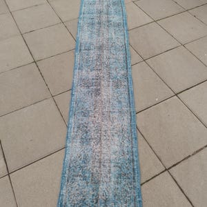 May include: A long, rectangular rug with a faded, distressed appearance. The rug features a color palette of blues, grays, and white, with a central design and a border. The rug is likely made of woven material and is suitable for a hallway or entryway.
