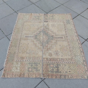 May include: A rectangular beige rug with a central geometric design in grey and brown. The rug has a border with repeating patterns in shades of green, pink, and brown. The rug is placed on a grey tiled surface.
