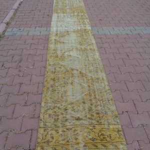 May include: A long, narrow, yellow and white patterned rug with fringe on the ends. The rug is lying on a brick walkway.