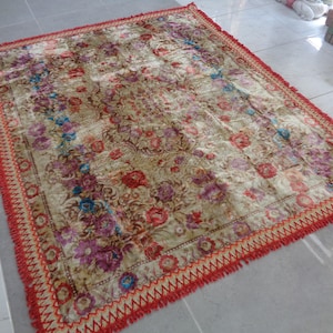 Puede incluir: Una alfombra vintage con estampado floral y borde de flecos rojo y amarillo. La alfombra tiene un fondo crema con diseños florales rojos, rosas, azules y amarillos.