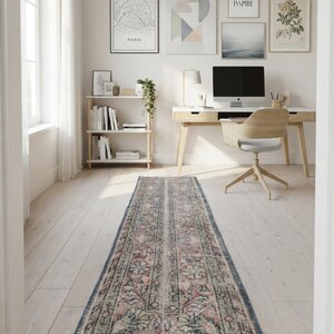 May include: A long, rectangular runner rug featuring a faded, intricate floral pattern in muted shades of dusty pink, sage green, and slate blue. The rug has a distressed texture with a fringed edge, centered on a light-colored wooden floor.