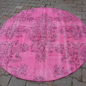 May include: A round pink rug with a faded floral pattern in a darker shade of pink. The rug has a distressed look and is made of a soft, woven material.