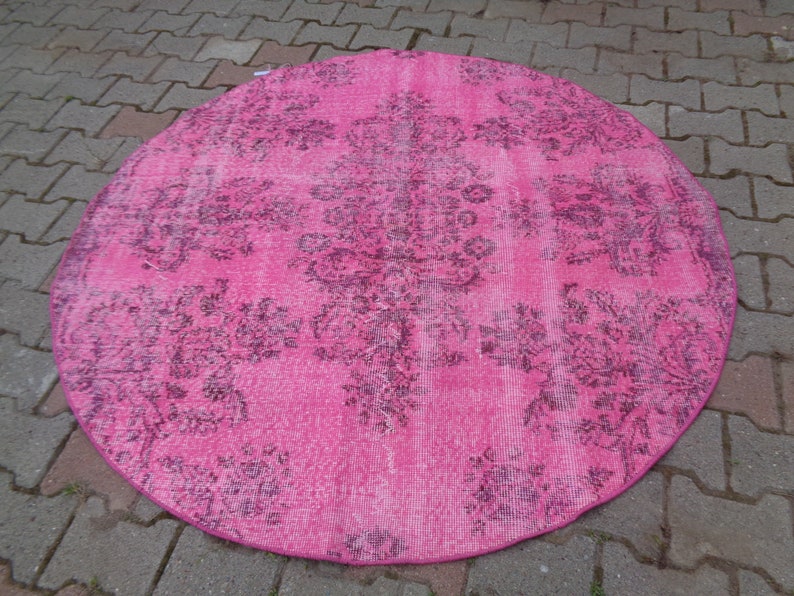 May include: A round pink rug with a faded floral pattern in a darker shade of pink. The rug has a distressed look and is made of a soft, woven material.
