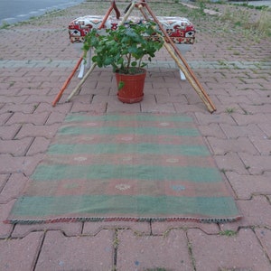 May include: A rectangular rug with horizontal stripes in shades of green and red. The rug has a fringed edge and small decorative patterns within the stripes. The rug is placed on a brick surface.