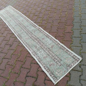 May include: A rectangular, narrow rug with a faded, vintage appearance. The rug features a central field of pale green with repeating floral and geometric patterns in muted shades of brown and cream. The rug's edges are bordered with a cream-colored fringe.