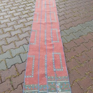 May include: A long, rectangular rug with a coral-colored background. The rug features a repeating pattern of blue vertical lines and geometric shapes. The border has a teal and green design. The rug is likely made of woven fabric.