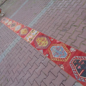 May include: A long, narrow red rug with a colorful geometric pattern. The rug is laid out on a brick walkway.