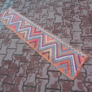 Puede incluir: Una alfombra larga y estrecha con un patrón geométrico de zigzags en tonos de rosa, azul, blanco y negro sobre un fondo melocotón.