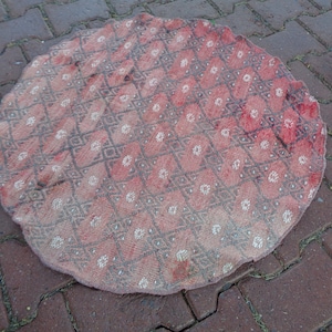 Puede incluir: Una alfombra redonda de color rosa y blanco con un diseño geométrico. La alfombra está hecha de tejido tejido y tiene un borde con flecos.