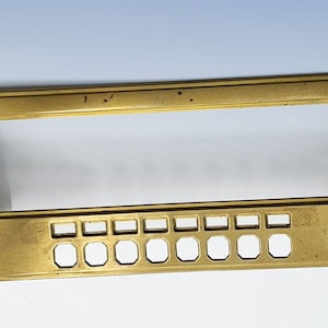 May include: A rectangular brass panel with a large central opening, smaller rectangular cutouts along the bottom, and two white and black accents on each end. The panel has a vintage or industrial aesthetic.