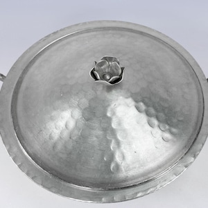 May include: A silver-colored, round serving dish with a lid. The lid features a flower-shaped handle and a textured, hammered surface. The dish has two handles and is likely made of metal. The overall design is vintage.