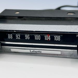 May include: A vintage Lafayette car radio receiver featuring a rectangular black and silver faceplate with a horizontal tuning dial marked 88 to 108, a circular tuning knob on the left, a push button on the right, and a black metal mounting bracket.