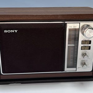 以下が含まれることがあります： ダークブラウンの木目調仕上げのキャビネットと黒いスピーカーグリルを備えたソニーの卓上ラジオ。フロントパネルには、垂直チューニングダイヤル、大きなチューニングノブ、トーンと電源ボリューム用の小さなノブを備えたシルバーのコントロールセクションがあります。