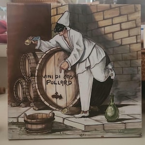 May include: A ceramic tile featuring a Harlequin figure in white attire and a black mask, leaning on a large wine barrel. The barrel is labeled "VINI DI CASA POLLARD." Additional barrels, a wooden bucket, and a green bottle are also depicted.