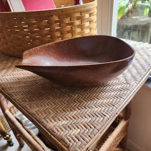 May include: A brown ceramic serving bowl with a unique, slightly curved shape. The bowl is sitting on a woven wicker surface.