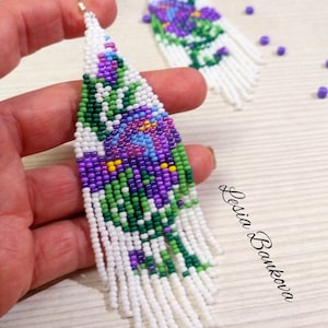 May include: Handmade beaded earrings with a floral design. The earrings feature a white fringe and a pattern of purple, green, blue, and yellow beads. The earrings are held by a hand, with a second earring visible in the background.