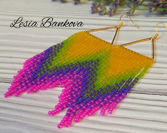 Op de afbeelding: Een paar lange, geperde oorbellen met een geometrisch, driehoekig ontwerp. De oorbellen zijn gemaakt van gele, groene, paarse en roze kralen. De oorbellen hangen aan gouden haakjes.