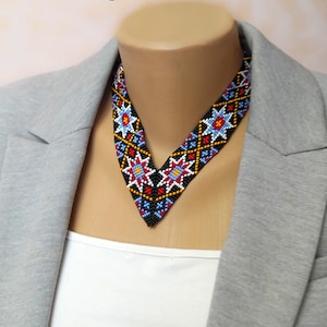 May include: A V-shaped beaded necklace with a black background and colourful geometric patterns. The necklace features red, blue, yellow, and white beads.