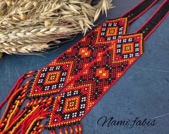 May include: A beaded necklace with a geometric pattern in red, black, orange, and white. The necklace has a black strap and beaded fringe. The text "Nami fabis" is visible in the image.