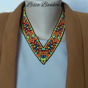 May include: A V-shaped beaded necklace with a geometric pattern in shades of orange, yellow, blue, and black. The necklace is on a mannequin wearing a brown jacket and a light blue shirt.