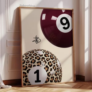 May include: A framed print featuring two billiard balls. One is a deep burgundy with the number 9, the other is a leopard print with the number 1. The print has a gold frame and a cream-coloured background.
