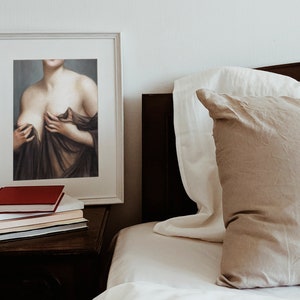 May include: A framed painting of a woman's torso, with her hands holding the fabric of a brown dress. The painting is hanging on a white wall above a bedside table with books.