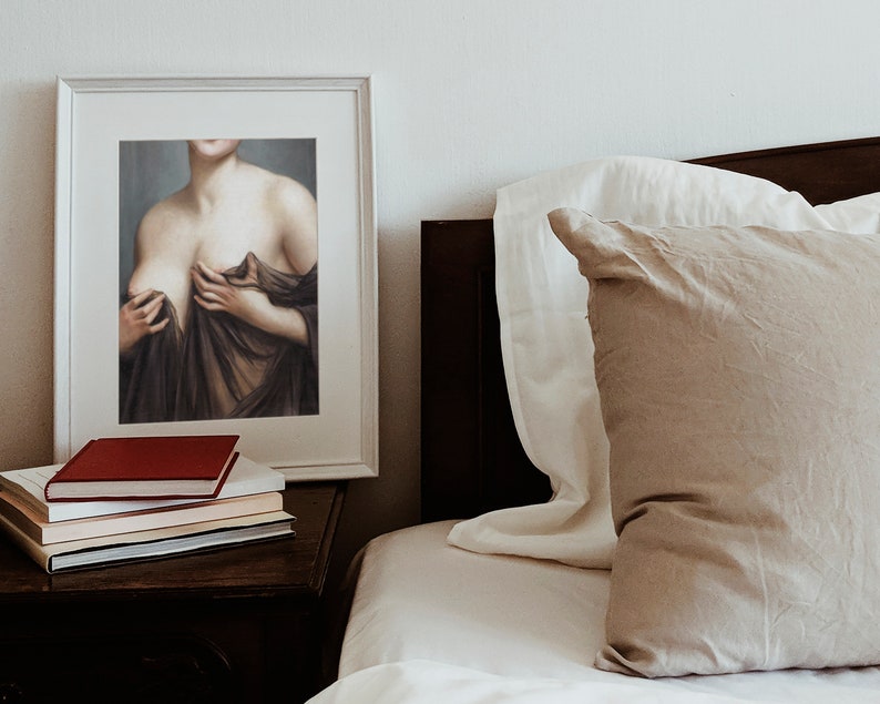 May include: A framed painting of a woman's torso, with her hands holding the fabric of a brown dress. The painting is hanging on a white wall above a bedside table with books.