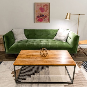 May include: A green velvet sofa with two pillows sits in front of a wooden coffee table with a geometric metal sculpture on top. The sofa is in front of a white wall with a framed floral print hanging above it.