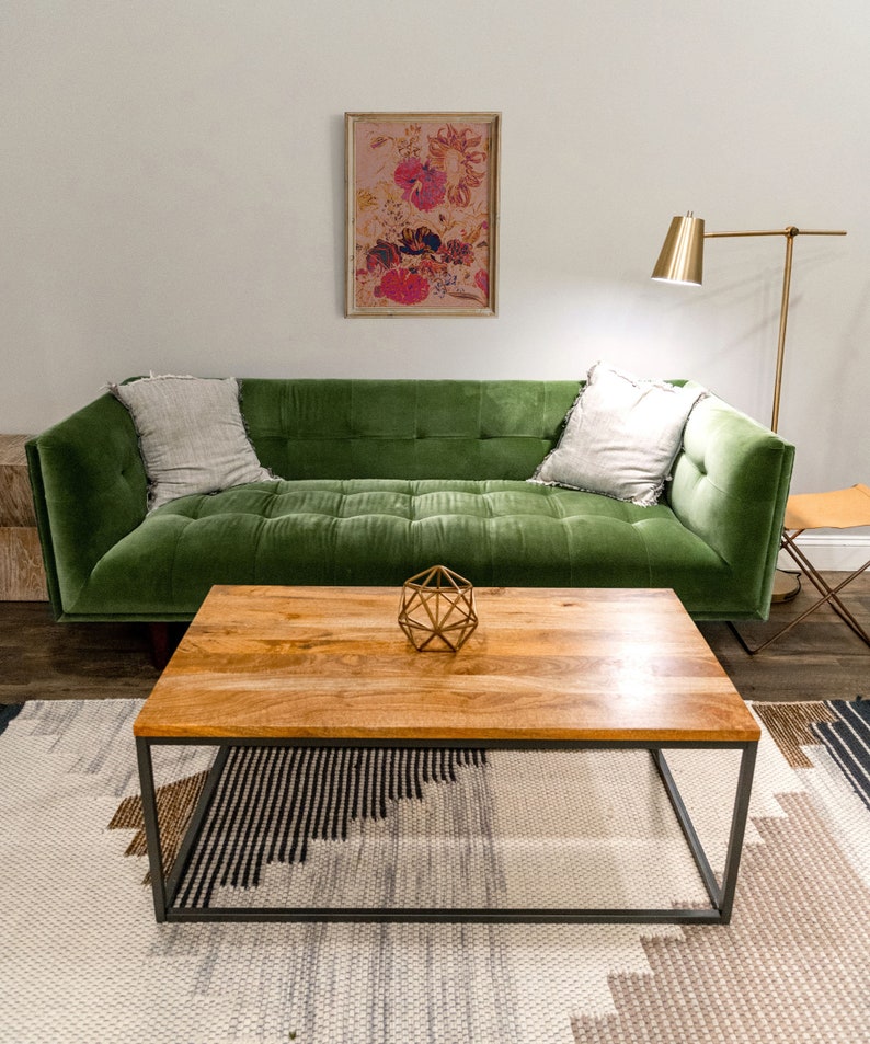 May include: A green velvet sofa with two pillows sits in front of a wooden coffee table with a geometric metal sculpture on top. The sofa is in front of a white wall with a framed floral print hanging above it.