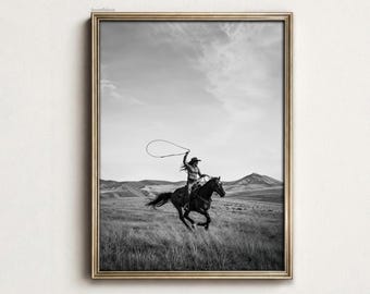 May include: A black and white framed wall art print depicting a rider on horseback galloping across an open field. The rider wears a cowboy hat and holds a lasso above their head. The background features rolling hills under a cloudy sky.