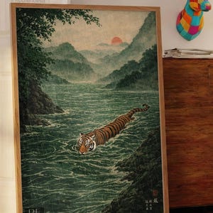 May include: Framed art print of a tiger swimming in a river, inspired by Japanese art. The artwork shows a tiger with orange and black stripes in a green river, with mountains and a sunset in the background. The frame is made of light brown wood.