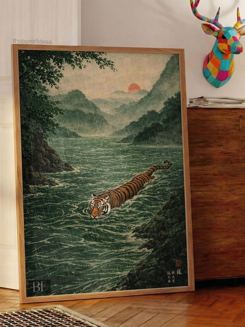 May include: Framed art print of a tiger swimming in a river, inspired by Japanese art. The artwork shows a tiger with orange and black stripes in a green river, with mountains and a sunset in the background. The frame is made of light brown wood.