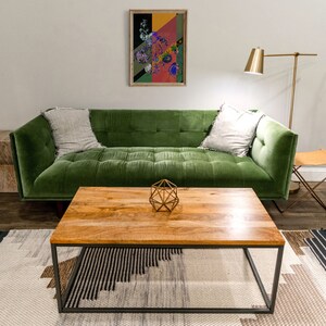 May include: A green velvet sofa with two pillows sits in front of a wooden coffee table with a geometric metal sculpture on top. The sofa is in a living room with a white wall and a floor lamp.