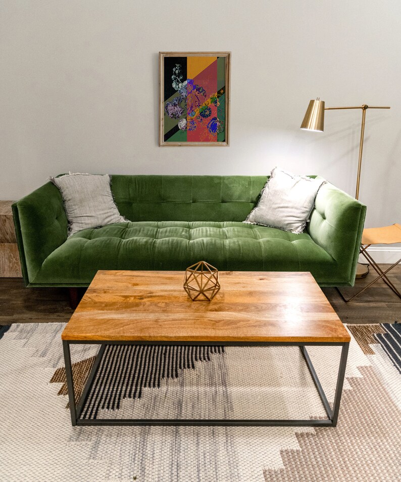 May include: A green velvet sofa with two pillows sits in front of a wooden coffee table with a geometric metal sculpture on top. The sofa is in a living room with a white wall and a floor lamp.