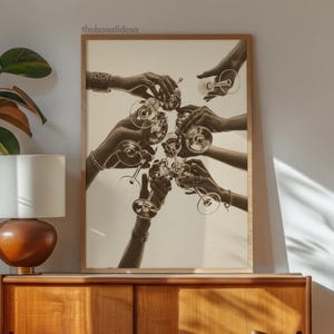 May include: Sepia-toned print of hands toasting with champagne glasses. The image features multiple hands holding and clinking glasses together, creating a celebratory atmosphere. The artwork is framed in a light brown wooden frame.