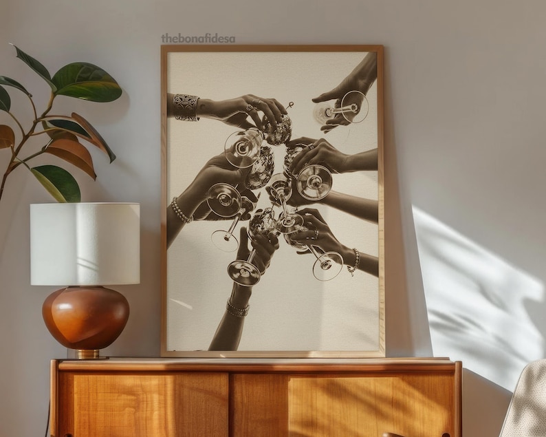 May include: Sepia-toned print of hands toasting with champagne glasses. The image features multiple hands holding and clinking glasses together, creating a celebratory atmosphere. The artwork is framed in a light brown wooden frame.