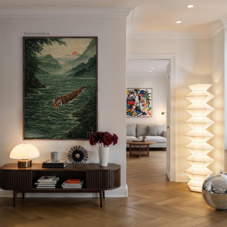 May include: A living room interior with a dark wood console table, a lamp, books, and decorative items. A large framed artwork shows a tiger swimming in a river. A tall, white, layered floor lamp and a disco ball are also visible.