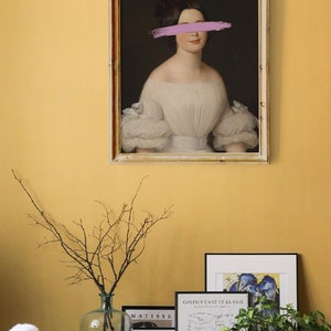 May include: A framed portrait of a woman in a white dress with a pink brushstroke covering her eyes. The portrait is hanging on a yellow wall with other framed artwork and a vase with branches.
