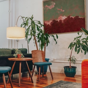 May include: A living room interior with a green and red abstract painting on the wall, a green potted plant, a wooden chair with a blue cushion, a small round wooden table, a green sofa, and a cream-colored lamp.